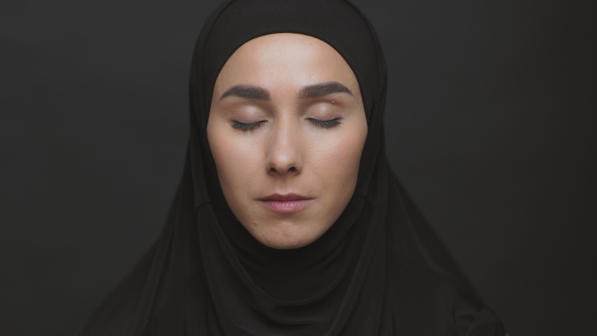 Female rights discrimination in muslim world. Close up portrait of young serious middle eastern woman wearing black hijab opening eyes and looking to camera, dark background | Shutterstock HD Video #1085182625