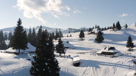 Aerial Raising Up, Tilting Down Shot of Alpine, Wooden, Holiday Village, Small houses, huts, Chalets Covered in Snow Standing On Peak of Hill With Spruce Forest, Hills and Alps in The Background.
