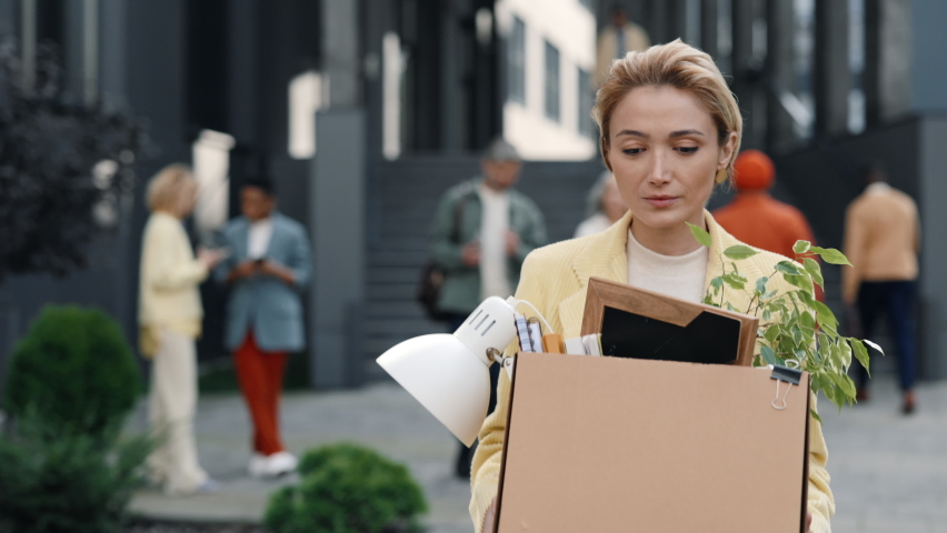 Young caucasian woman carrying paper box with personal stuff while leaving office center. Multicultural business people walking on background.