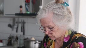 Elderly woman with gray hair wearing glasses in home kitchen, blurred background. Grandma reads the message carefully.