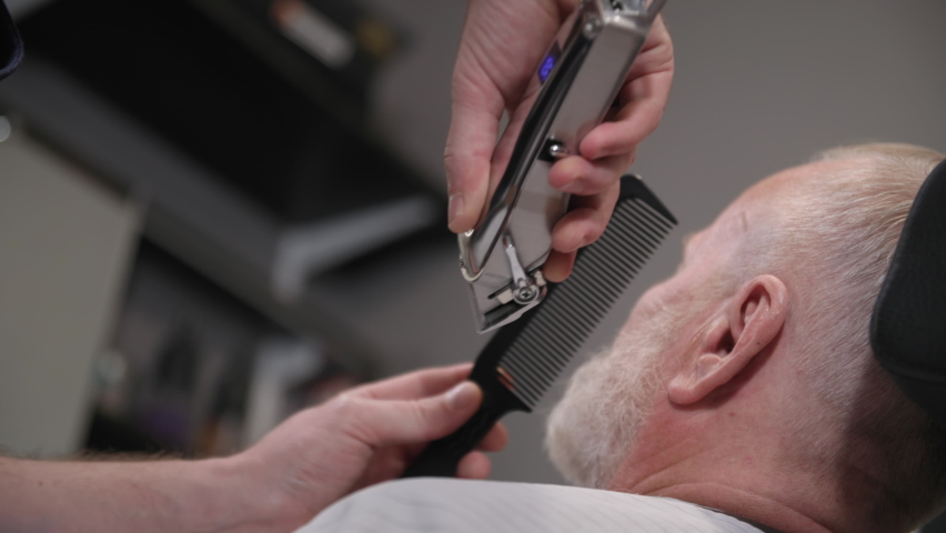 stylish male barber doing haircuts and styling to an elderly man with a beard using a trimmer and comb in a men