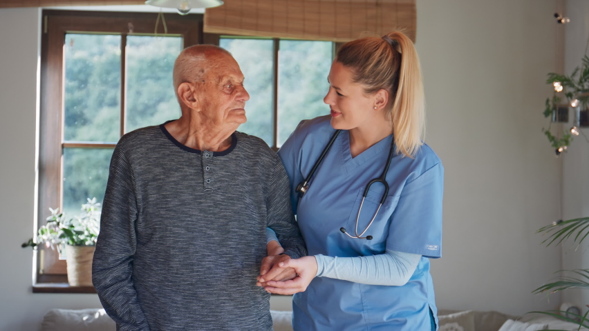 Young female healthcare worker visiting senior man indoors at home, holding and walking with patient. Royalty-Free Stock Footage #1085194460