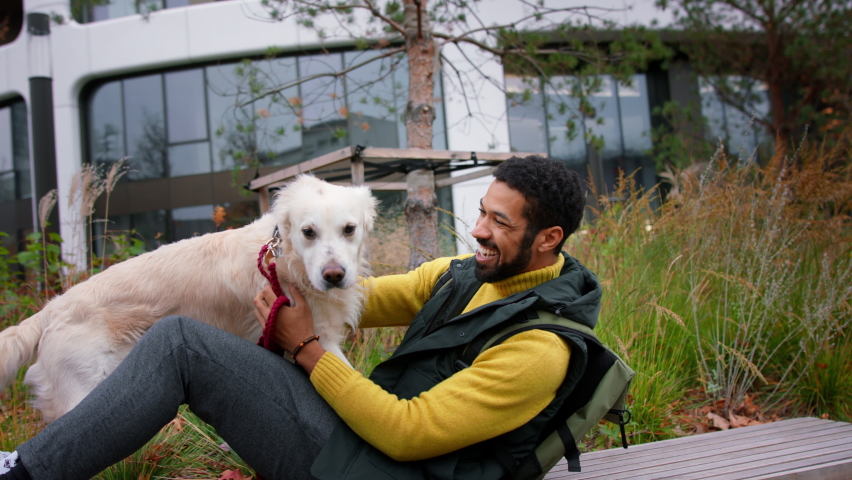Happy young man sitting and stroking his dog during walk outdoors in city in autumn.