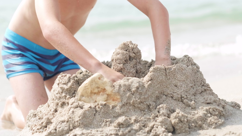 close up hands little blonde boy playing with sand on beach ocean sea. Child building sand castle house. Family summer holidays and trips to warm countries