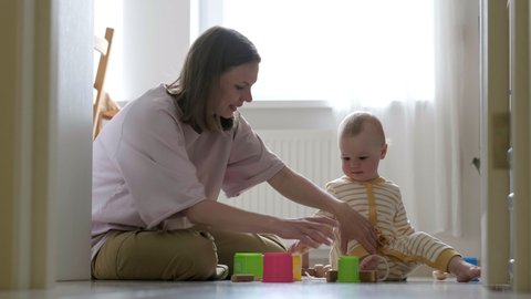 Little baby girl and mommy play with colorful toys at home sitting on floor. Mother and daughter laughing having fun together.