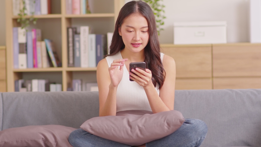 Millennial asian young woman looking mobile phone laughing with good news or discount voucher for shopping online at home.Happy and cheerful woman looking on cellphone app read message feel excited Royalty-Free Stock Footage #1085196917