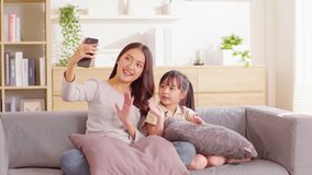 Asian young mother and her daughter have video call conference with family having fun together.Happiness Mom and little girl looking at mobile phone and waving hand with video chat to grandmother 