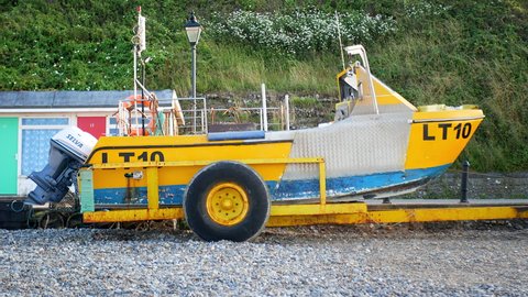 Cromer, Norfolk, Circa September 2021. Yellow and blue crab and lobster fishing boat on pebble beach with beach huts and grass cliff behind.