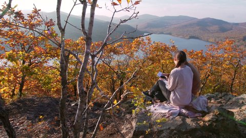 Back of couple sitting on the big stone on the mountain peak, drinking hot tea, reading a book and enjoying the landscape of autumn forest and blue river far away under the bright sun