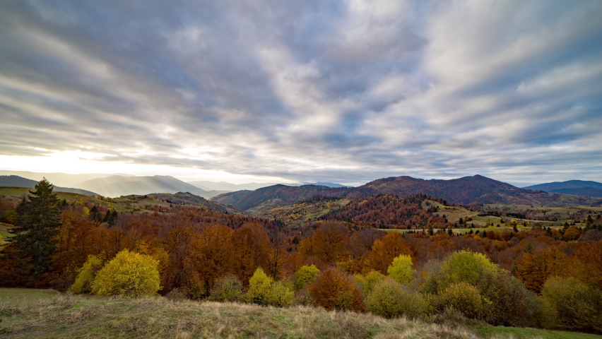 Colorful autumn trees with orange and yellow leaves grow in highland against big mountains and hills under floating heavy clouds 8K video time-lapse