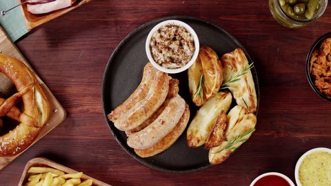 Putting plate with smoked sasuages and fried potatoes top view, pickled cucumbers, bratwursts and fresh pretzels on table. Traditional German Cuisine. Composition of Cooked National Czech Food. 