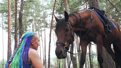 lgbt. rainbow. equestrian riding. rider girl, horsewoman, with rainbow hair dreadlocks, is treating her horse with goodies, while walking through the forest