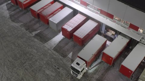 Aerial view of a semi-trailer truck drives up to a warehouse ramp for loading and unloading goods in a logistics park at winter night