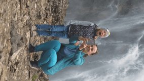 Vertical Video. Young Woman with Little Daughter Taking Selfie with Smartphone by a Mountain Waterfall. Ecotourism and Travel Lifestyle Concept