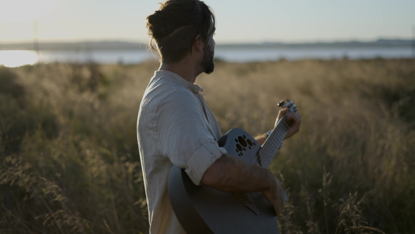Man Guitarist Standing in the grassy field in Spain and playing chords on black acoustic guitar against beautiful sun light - slow-motion profile view