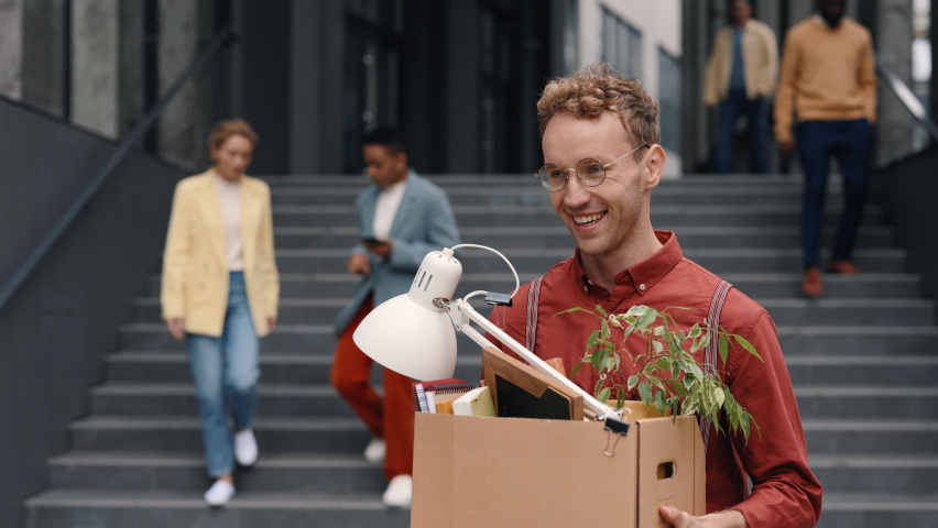 Caucasian man in eyeglasses and stylish formal wear carrying box with office stuff outdoors. Happy young guy with curly hair leaving office center after quitting job. Royalty-Free Stock Footage #1085222693