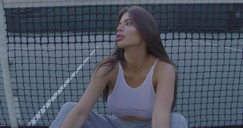 Portrait of a sporty sexy girl with big tits sitting near the net on a tennis court in a white bikini in the city center. Red camera, slow motion. A model with a tail enjoys life