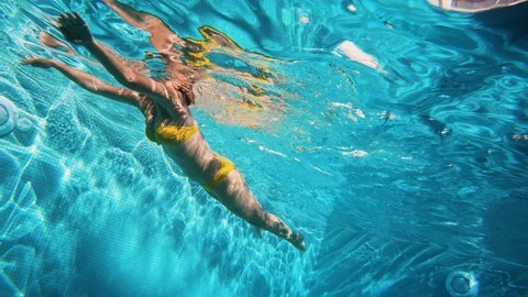 4K Cinematic slow-motion underwater footage of a woman in a yellow swimsuit swims in clear blue water in the pool