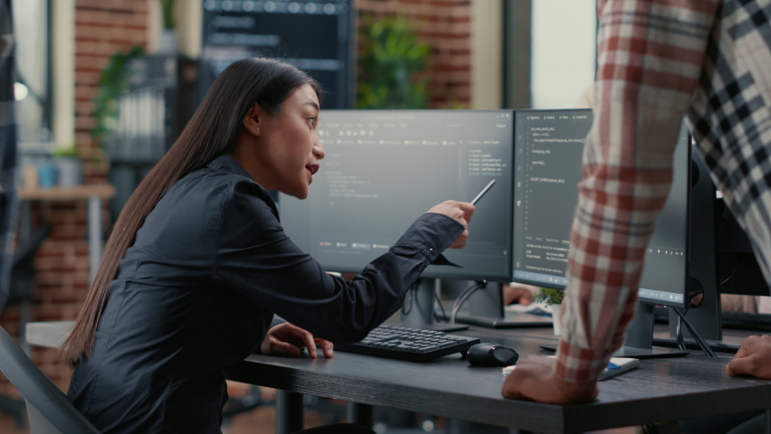 Database developer pointing pencil at computer screen with software compiling code explaining source algorithm to coworker standing next to desk. System engineers discussing ai team project. Royalty-Free Stock Footage #1085228792