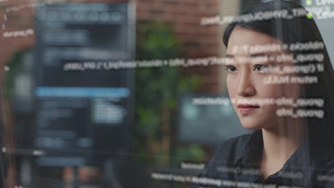 Portrait of focused asian software developer through vfx of floating programming code writing and smiling sitting at desk. App developer looking at hologram of machine learning data algorithm.