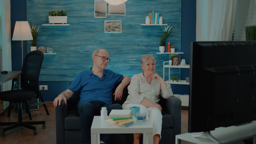 Retired couple watching comedy movie on television at home, enjoying free time together. Old man and woman sitting on couch and looking at film on tv for entertainment. Media channel Royalty-Free Stock Footage #1085228936