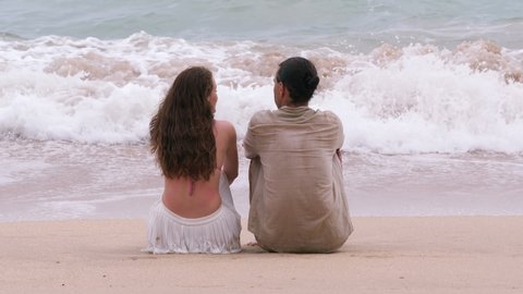 Couple sit on sand, look to breaking waves, telephoto view from back, flat perspective. Woman and man spend time together at tropical sea beach, backpacker vacation concept