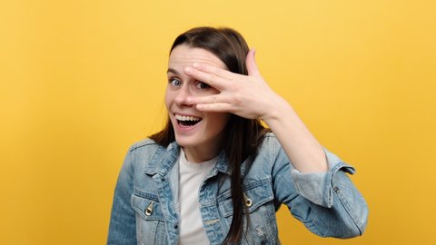 Portrait of young caucasian woman looks through fingers, peeks with curious face, having suspicious, wears denim jacket, posing isolated over yellow color background in studio. Watches secrets concept