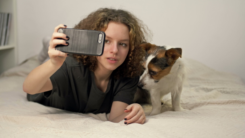 Beautifu teenage girl ltake a photo selfie bu mobile phone with her cute dog in cozy bedroom.