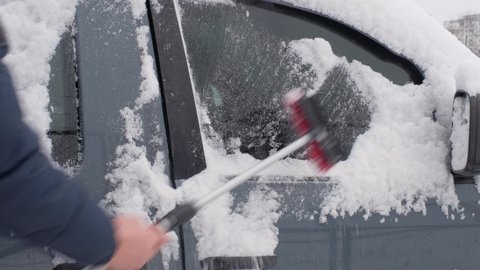 Man brushing fresh snow from his car with a brush. The streets are covered with snow and drifts. Winter season.