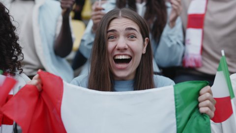 Young woman screaming emotionally while supporting italian team during soccer game. Female fan with flag in hands sitting on bleachers and looking at camera.