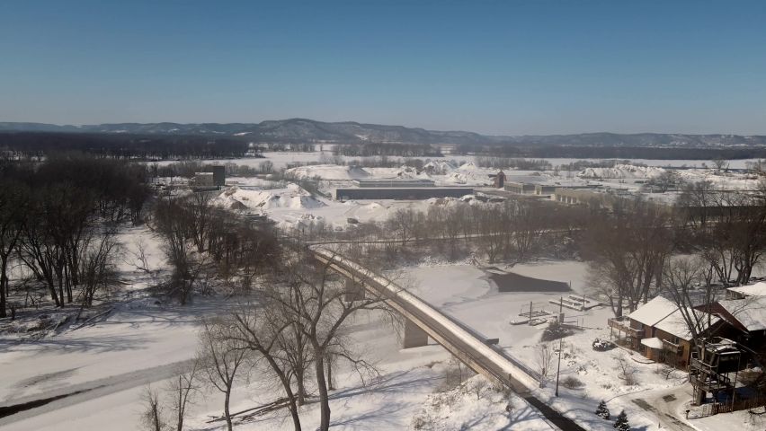 Snow covered landscape with icy river and mountains in the background with brilliant blue sky, Winter in Wisconsin. Aerial view over river with bridge and residential neighborhood seen. 