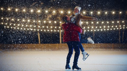 Romantic Winter Snowy Evening: Ice Skating Couple Meeton on Ice Rink and Have Fun. Pair Skating Boyfrined Lifts His Beautiful Girlfriend and Spins. Love, Dance, Embrace, Figure Skate. Wide Slow Motion