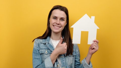 Cute young caucasian woman holding paper house and smiling to camera, advertising real estate agency, dressed in denim jacket, posing isolated over yellow studio background wall. Home purchase concept