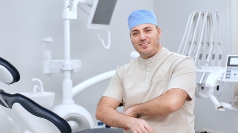 Portrait successful male dentist in uniform smiling posing with crossed hands medical clinic operating room. Happy stomatologist orthodontist orthopedist at professional teeth care treatment equipment