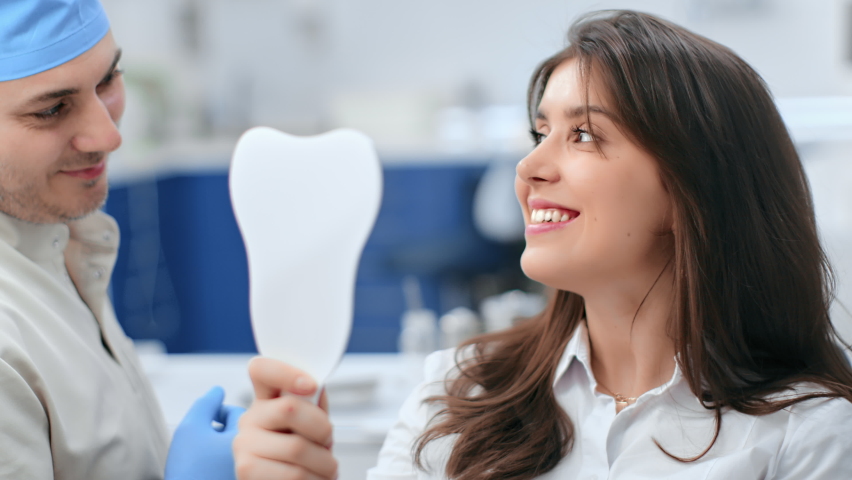 Happy woman looking at mirror enjoy healthy clean teeth beautiful smile after treatment at stomatology clinic. Pretty female patient and dentist doctor admiring result tooth whitening at dentistry  | Shutterstock HD Video #1085256593