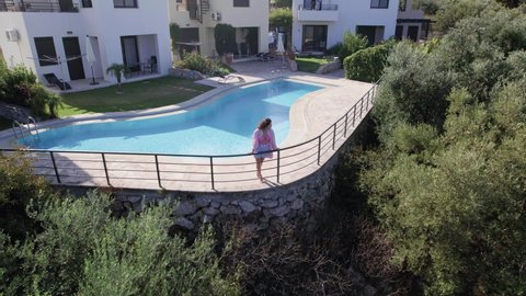 Drone footage of woman walk near swimming pool, and Modern Villas in scenic countryside. Luxurious Wealthy Neighborhood, Beautiful Homes in Crete Greece. Aerial Pan Out View.