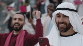 Positive bearded man in traditional sheikh clothes live streaming soccer game using modern smartphone. Happy fan standing on tribune with gadget in hands.