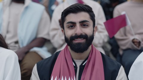 Portrait of happy muslim man with beard enjoying football match on big stadium. Young guy smiling and looking at camera with other fans around.