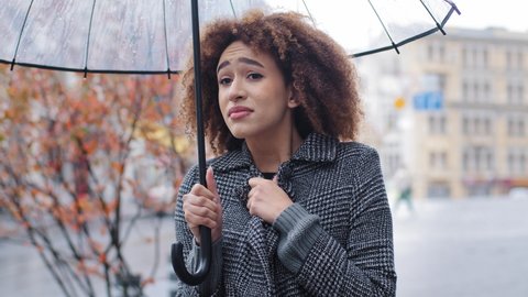 African American girl curly-haired sad woman in stylish coat stands in autumn on city street with transparent umbrella in rain suffers from cold winds low temperature waiting for taxi looking around