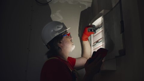 Electrician with phone flashlight fix electricity in fuse box at construction site. Realtime. 