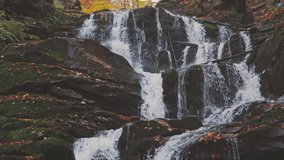 Slow-motion 4K zoom video waterfall stream falling down rocks and stones of mountain steep slope, forest with yellowed trees and terracotta fallen leaves on autumn day