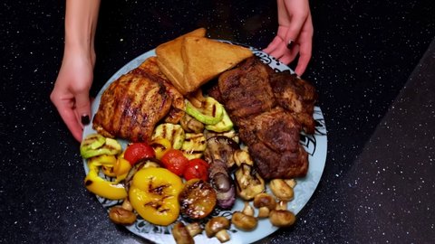 A housewife puts fried juicy meat steaks with spices and herbs and grilled vegetables on a plate. Juicy barbecue with grilled stripes is ready for a picnic celebration. Barbecue grill and crispy toast