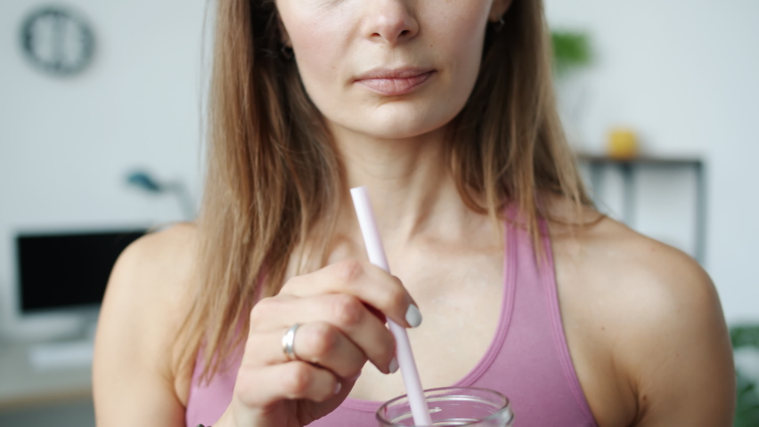 Close-up portrait of young sportswoman in bra drinking smoothie with straw and smiling at home. Healthy lifestyle and useful nutrition concept.