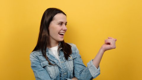 Portrait of young caucasian woman looking playfully and calling with one finger, making beckoning gesture, inviting to come, wearing denim jacket, posing isolated over yellow color background studio