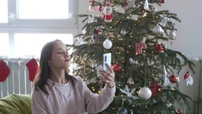 Beautiful Young Smiling Woman near Christmas Tree with Shining Lights at the Background Making Video Message or Selfie Concept of Holidays and New Year in Christmas decoration room.