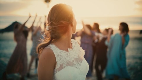Portrait of a Beautiful Bride in Pure White Wedding Dress Tossing Bridal Bouquet and Passing Good Fortune to Her Happy Multiethnic and Diverse Friends. Outdoors Venue on a Beach Near Sea or Ocean.