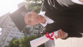 Handsome guy graduate taking selfie video in front of the camera he holding diploma and speaks something very excited in the college garden taking video horizontally. 4k