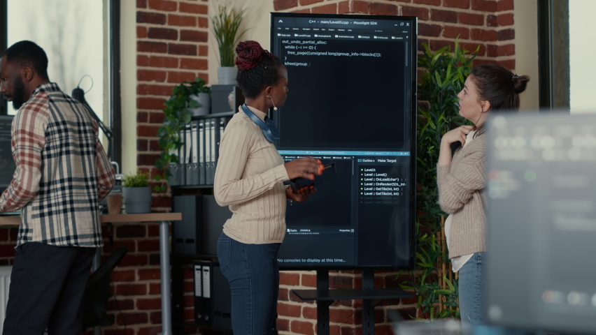 Programmer holding digital tablet analyzing code on wall screen tv while colleague is looking for errors pointing using remote control. Team of app developers collaborating on big data group project. Royalty-Free Stock Footage #1085297660