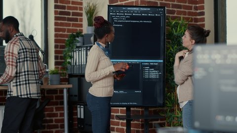 Programmer holding digital tablet analyzing code on wall screen tv while colleague is looking for errors pointing using remote control. Team of app developers collaborating on big data group project.