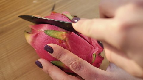Close-up of hands cutting a pitahaya fruit with a black knife on a wooden board.